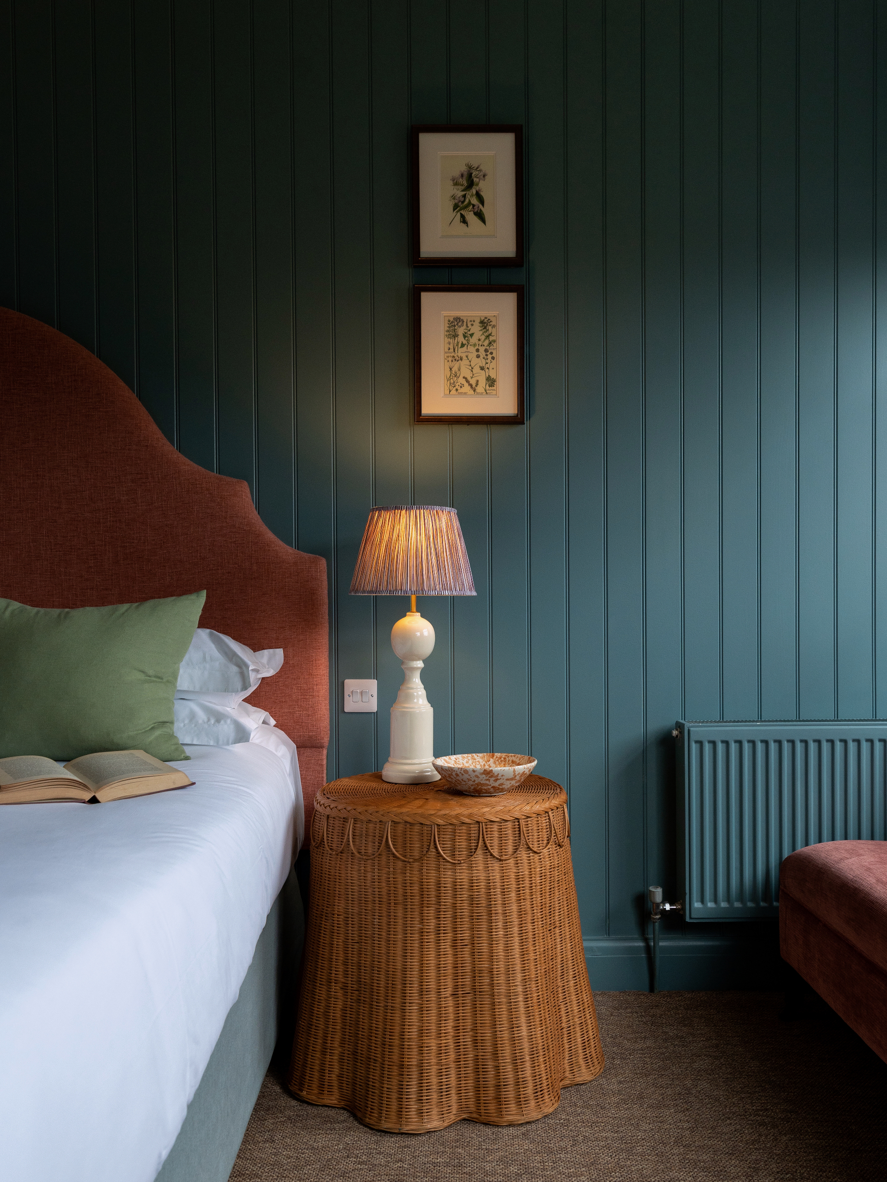 Cozy bedroom with dark blue paneled walls, headboard upholstered in paprika rust fabric and a caramel rattan side table