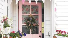 Spring front door ideas like this one are so pretty. Pictured is a pastel pink front door with a pink and blue wreath on it, bunting above it, and white, purple and pink flowers in pots in front of it