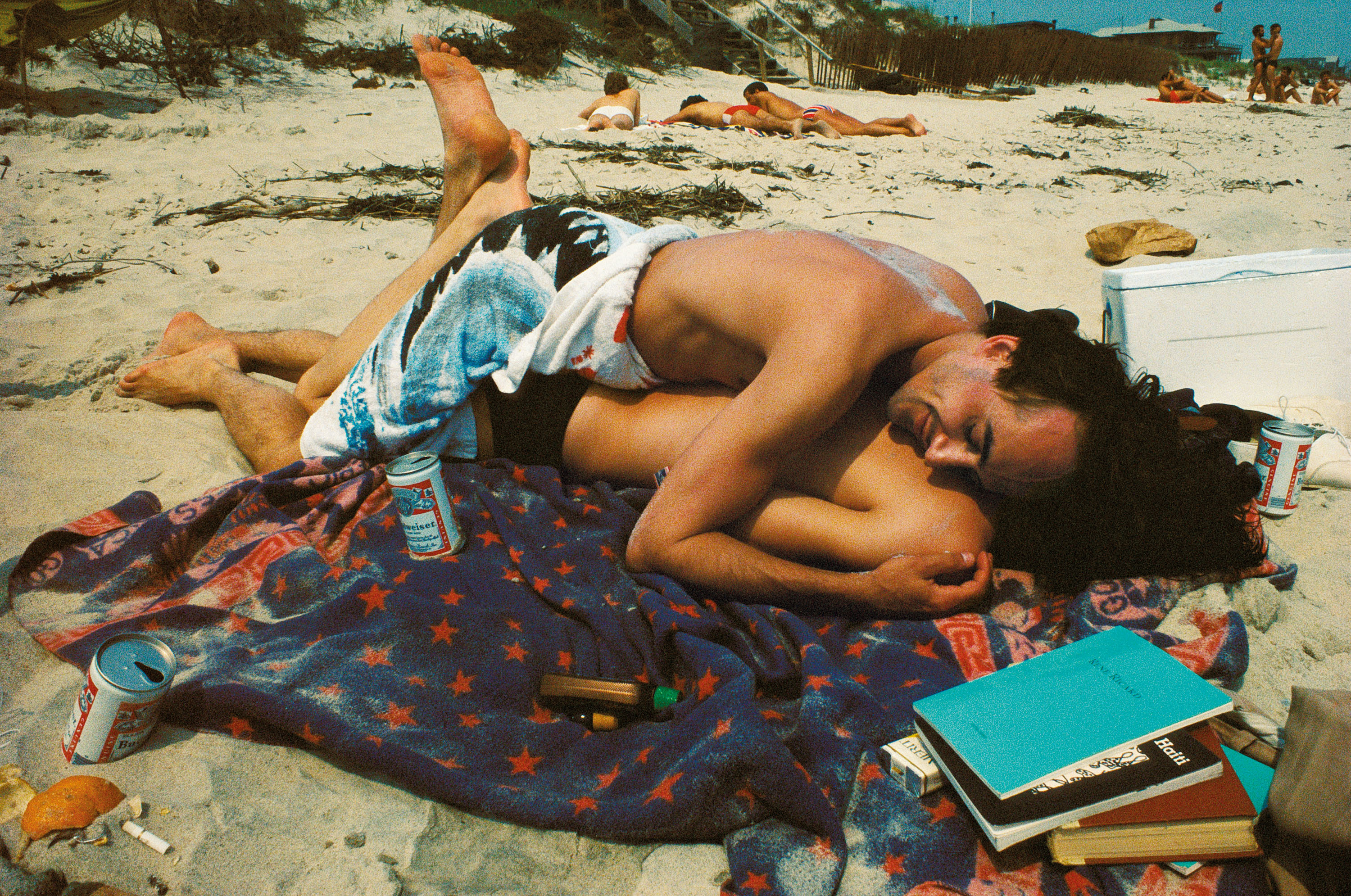 Two people embrace while lying on a star-patterned towel on a sandy beach, surrounded by books, drinks, and other beachgoers in the distance.