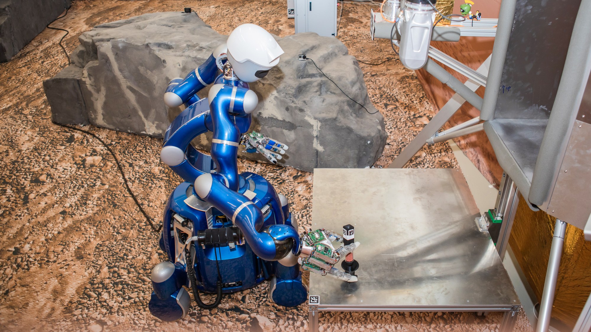 a humanoid robot on a mars-like landscape, beside a platform. the hand of the robot is outstretched to the platform and a sample is on top of it