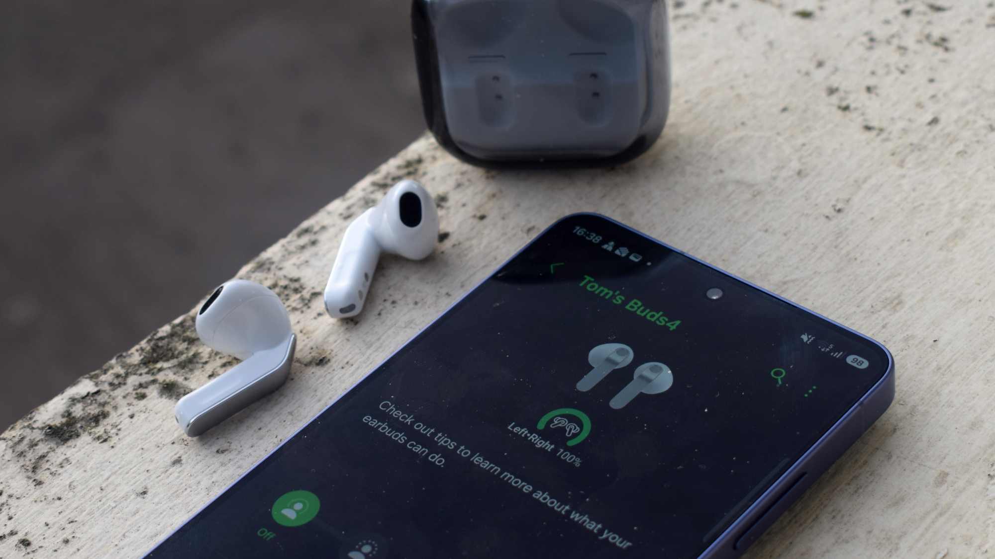 The Samsung Galaxy Buds 4 earbuds on a ledge, next to a Samsung phone showing the buds app.