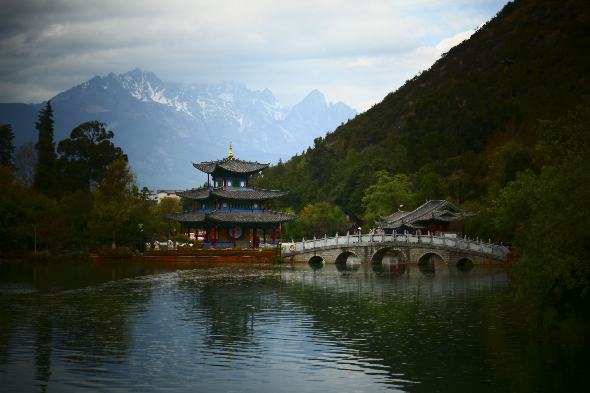 A Chinese temple by the side of a lake with an arched bridge leading up to it and both are reflected in the still water, in front of a snow covered mountain. A creative filter is applied to the image