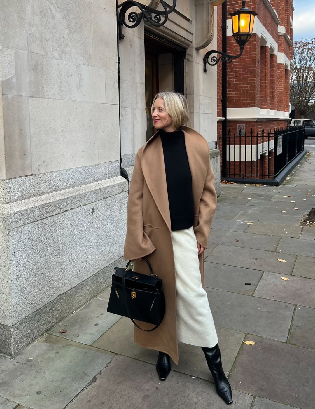A woman wears a white skirt, tan coat, and black boots