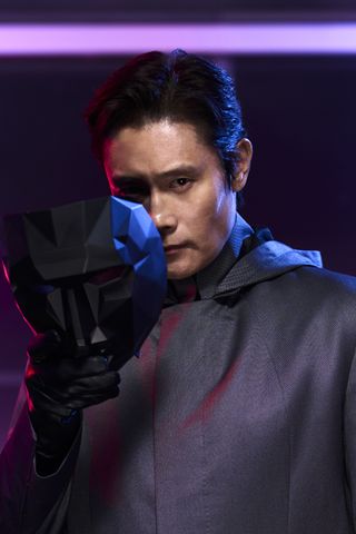 a man (lee byung-hun as the front man) holds his black mask partly covering his face as he looks at the camera