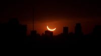 the sun appears as a thin crescent and glows a deep orange hue above silhouetted buildings it is low in the sky.