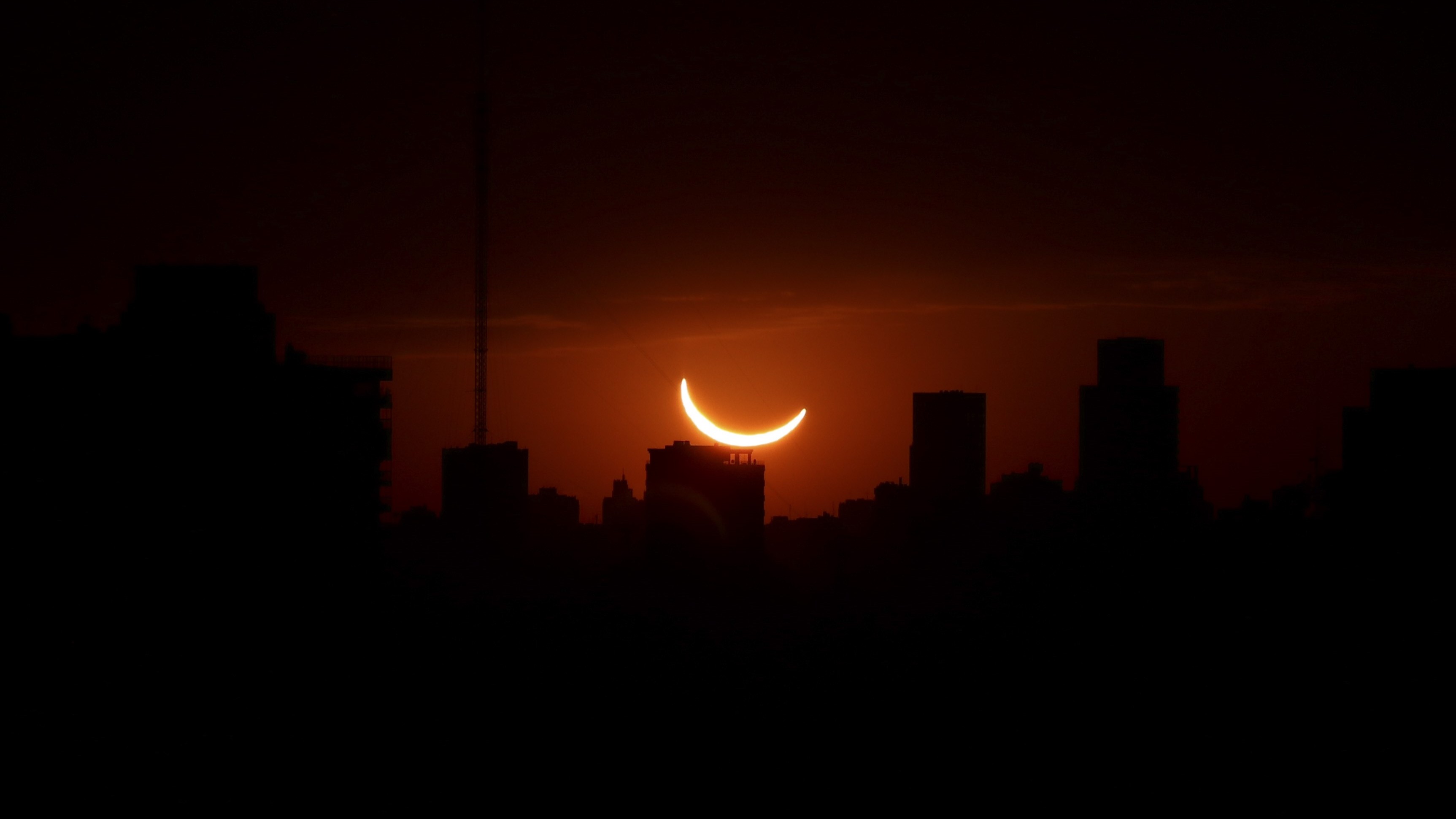 the sun appears as a thin crescent and glows a deep orange hue above silhouetted buildings it is low in the sky.