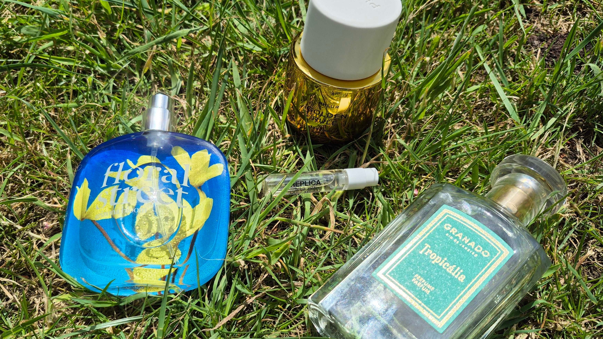 A small selection of the Perfumes that smell like suncream from this roundup, including Floral Street's Arizona Bloom, Phlur's Golden Rule, a small tester of Maison Margiela's REPLICA Beach Walk perfume and Granado's Tropicalia, all pictured on some green grass, in the sunshine.