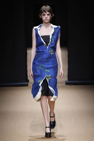 A female model wearing a sleeveless floral patterned blue dress and black shoes walking down a runway.