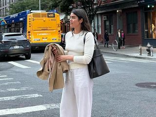 British influencer Anna Newton poses on a New York City street wearing an ivory ribbed long-sleeve tee, black Toteme Belted Leather Tote, a canvas jacket, and white linen pants