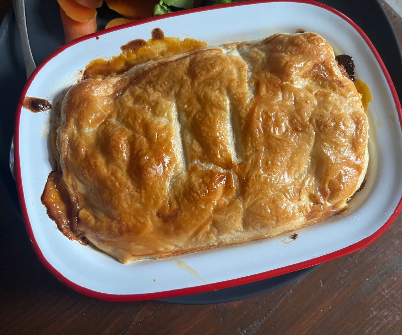 A white enamel dish with a red rim, with a puff pastry-topped pie inside.