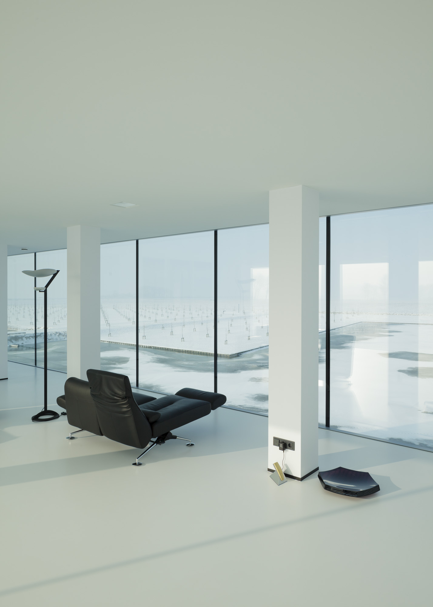 A bare, white room furnished with a black futon looks out onto a flat snowy landscape.