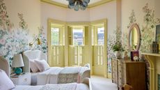 A large bedroom with traditional decor, twin beds, a butter yellow bay window and a floral wall mural.