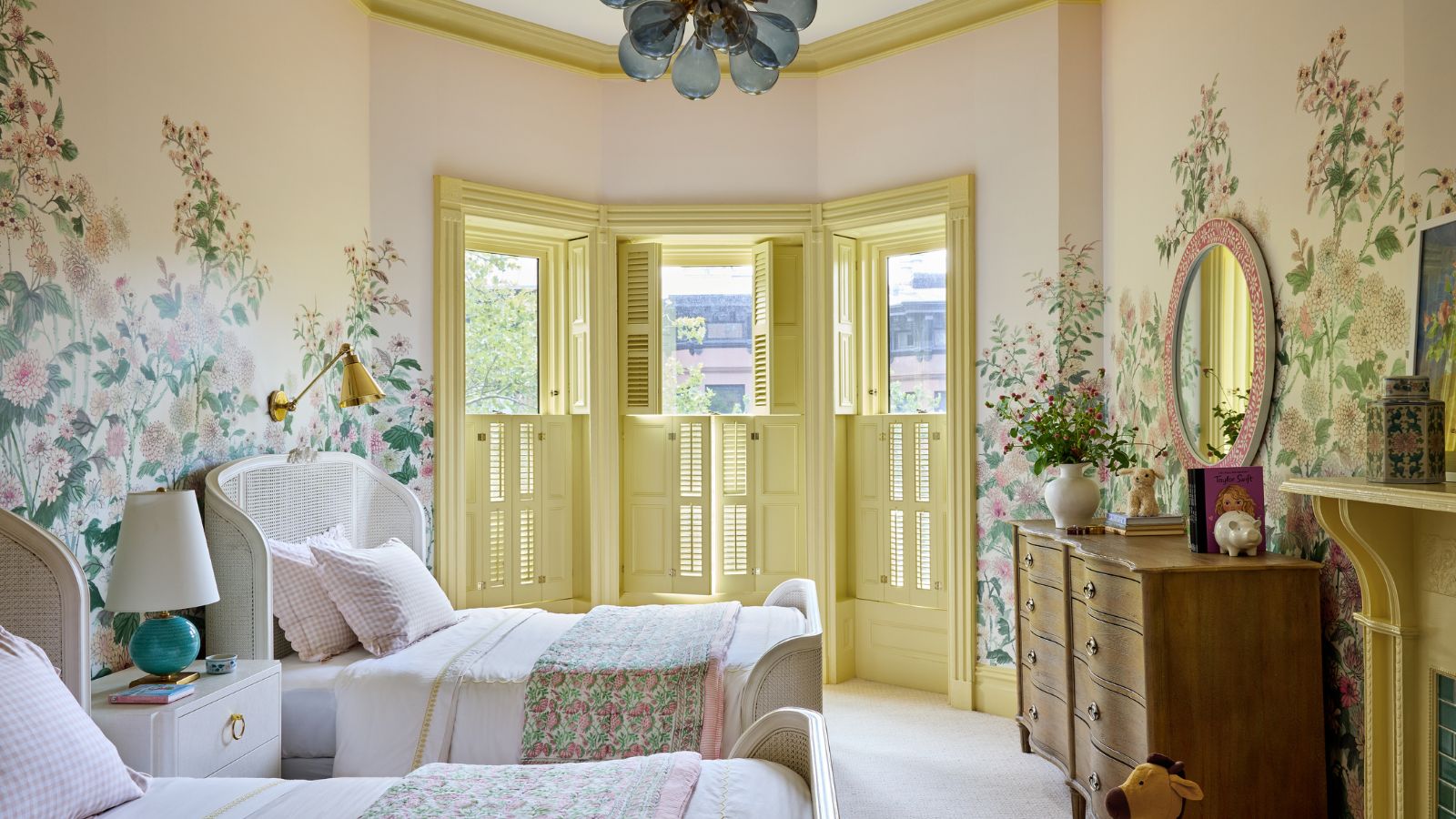 A large bedroom with traditional decor, twin beds, a butter yellow bay window and a floral wall mural. 