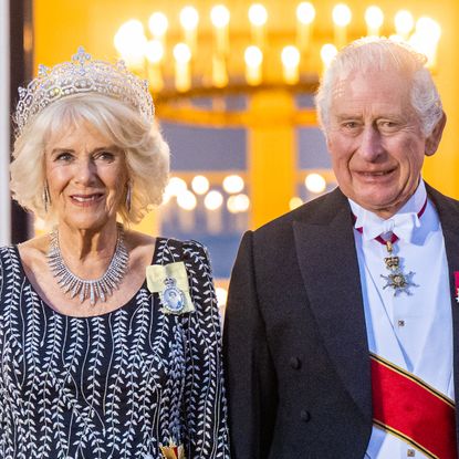 Queen Camilla wears a sparkling tiara and a black dress with a white floral design, while King Charles wears a formal suit with medals and a red sash