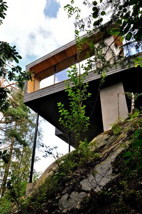 Alternative exterior view from below of the elevated Trekronekabin - a dark wood house that sits on rocks surrounded by trees under a blue cloudy sky