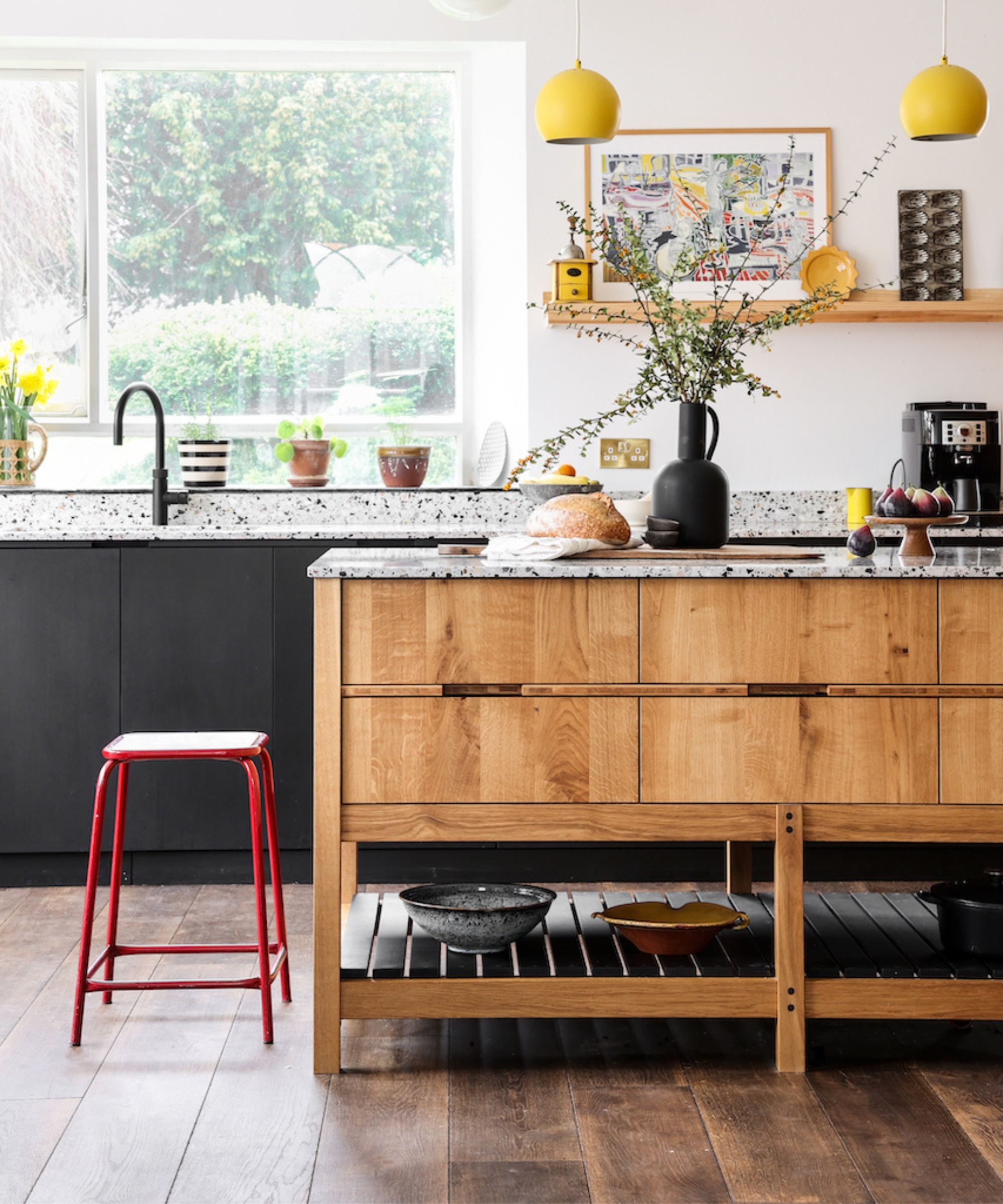Oak kitchen island with terrazzo top beside black units, red metal stool, yellow pendants and large window over sink