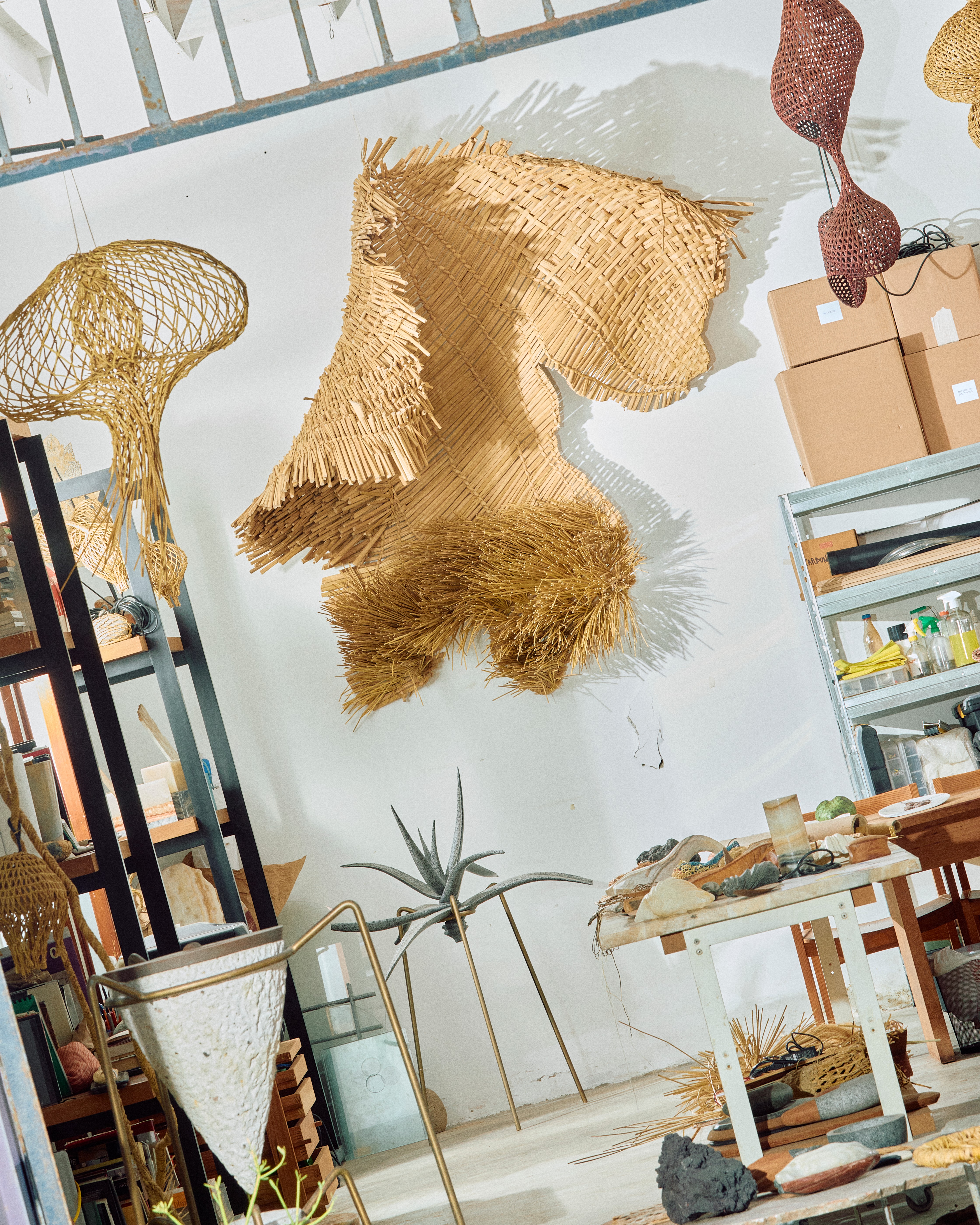Interiors of an artisan workshop filled with rattan, wood, stone, and ceramic sculptures and light, among which sit or stand its artisans.