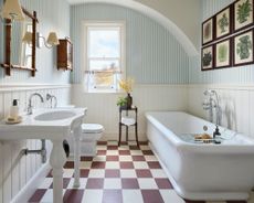 Can you paint a tile floor? Large bathroom with blue striped wallpaper, red and white chequerboard flooring, traditional white bath, sink and toilet, bamboo square mirror, wooden cabinet, two wall lamps beside mirror, botanical prints above bath with wooden frames