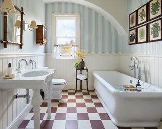 Large bathroom with blue striped wallpaper, red and white chequerboard flooring, traditional white bath, sink and toilet, bamboo square mirror, wooden cabinet, two wall lamps beside mirror, botanical prints above bath with wooden frames