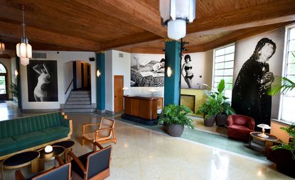 Freehand hotel in Miami. Light tiled floors, with wooden furniture with deep green and deep blue fabric. White walls, with blue columns and lots of plants throughout the space. Black & white photographs of models are hung on the wall. 