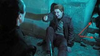 George Weasley sitting on the floor. Red arrows point to a wand a the leg of someone laying near him.