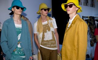 Three male models wearing looks from the Burberry Prorsum collection. One model is wearing a blue and white t-shirt, blue trousers and a blue cardigan. Another model is wearing a brown and white t-shirt with a castle design, brown trousers, a dual coloured scarf and is holding a briefcase. The third model is wearing a white shirt and mustard coloured jacket. All three models are wearing hats and sunglasses