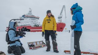 Three men stand on top of ice with a ship behind them