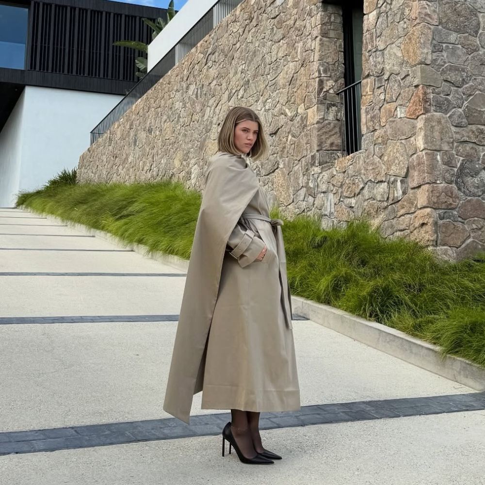 Sofia Richie poses for a photo outside wearing a long-line beige scarf coat with sheer tights and black pointed-toe heels.