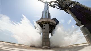 water sprays beneath a giant silver spacex starship rocket, which is sitting on a raised launch mount