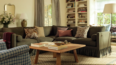 a cozy living room with a large green sectional, a large wood coffee table with decor on it, white painted walls, curtains, built-in bookshelves, and a floor lamp