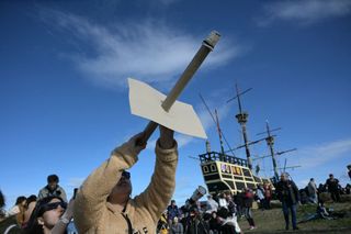 crowds gather to watch the eclipse, a person in the foreground is holding a long makeshift solar viewing device to protect their eyes while they look at the sun.