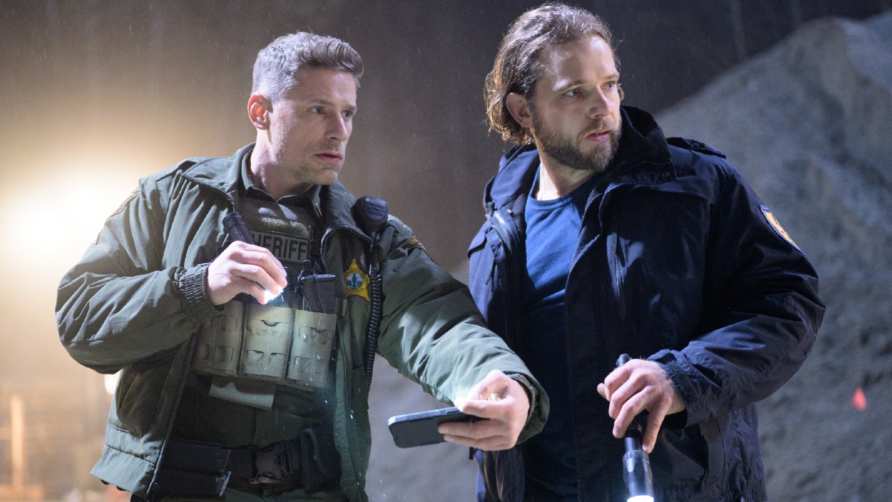 From left to right: Matt Lauria as Boone holding a flashlight in his left hand and a phone in his right hand while Max Thieriot stands next to him and holds a flashlight in his right hand. They're both looking to their right. 
