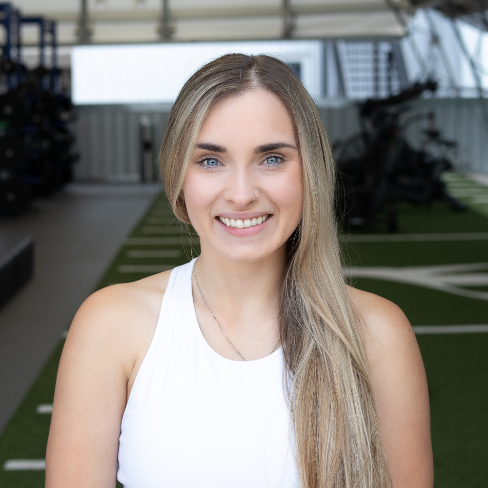 Headshot of smiling woman with long hair over one shoulder wearing white tank top with gym seen in background