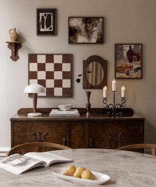 A dining area with five brown and white wall art prints and a sconce hanging on the wall, a dark brown wooden side table with candles and lamps, and a gray marbled dining table with a book and a bowl of pears