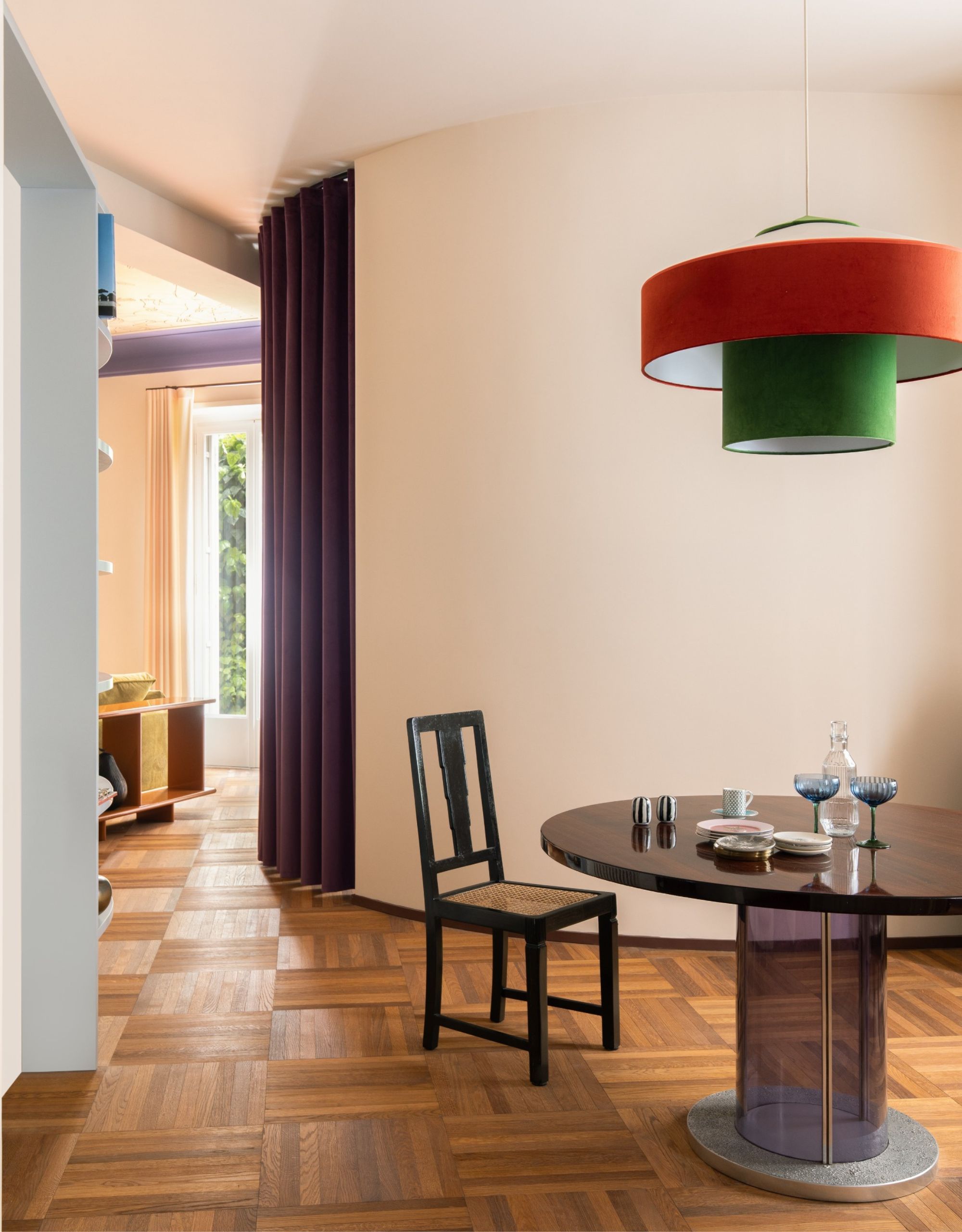 A dining room with parquet flooring, a round dining table, and colorful overhead ceiling light with a curved wall with a central purple curtain around it