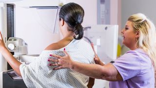 a black woman with short straightened hair wears a hospital gown and is facing away from the camera. A white woman wearing scrubs is helping position the patient for a mammogram