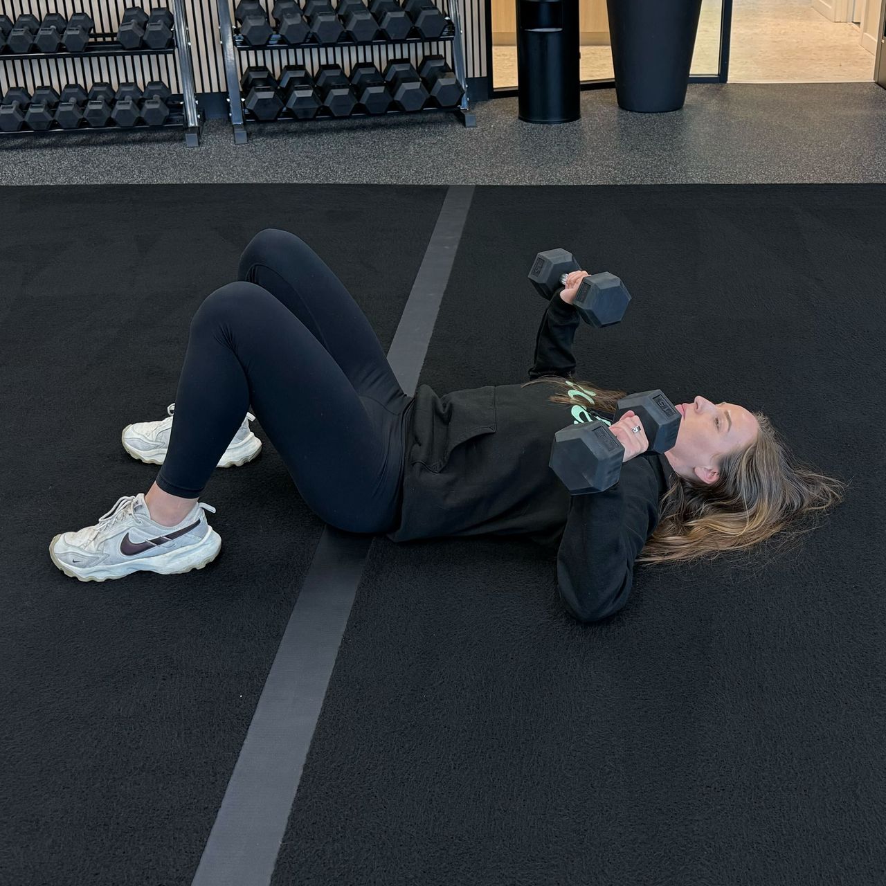 Felicia Hernandez, a personal trainer for Eden Health Club, performs a dumbbell chest press on the floor. She is lying with her knees bent and feet flat on the floor, elbows bent and dumbbells close to her shoulders.