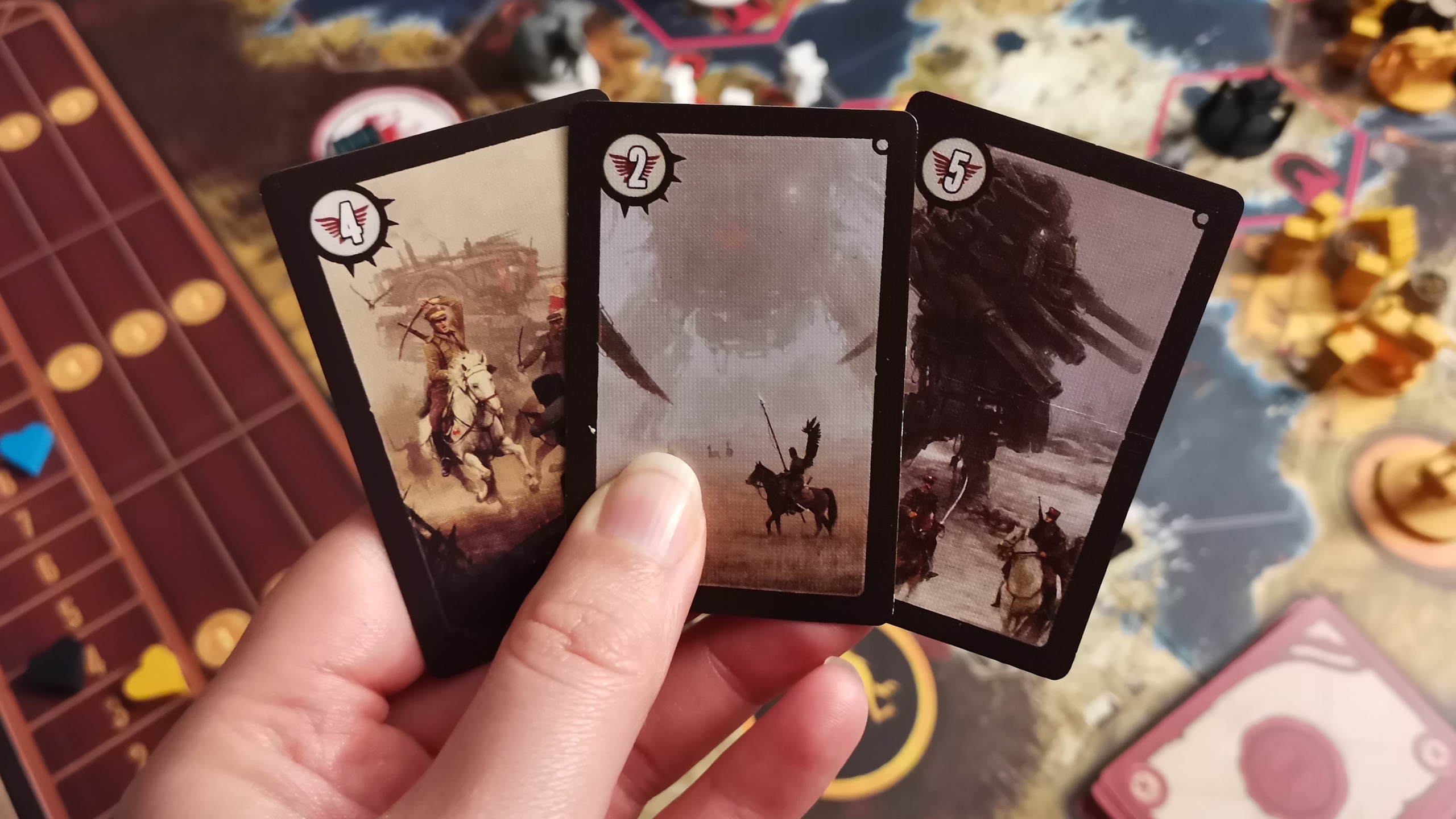 A hand holds Scythe cards, with the board visible in the background