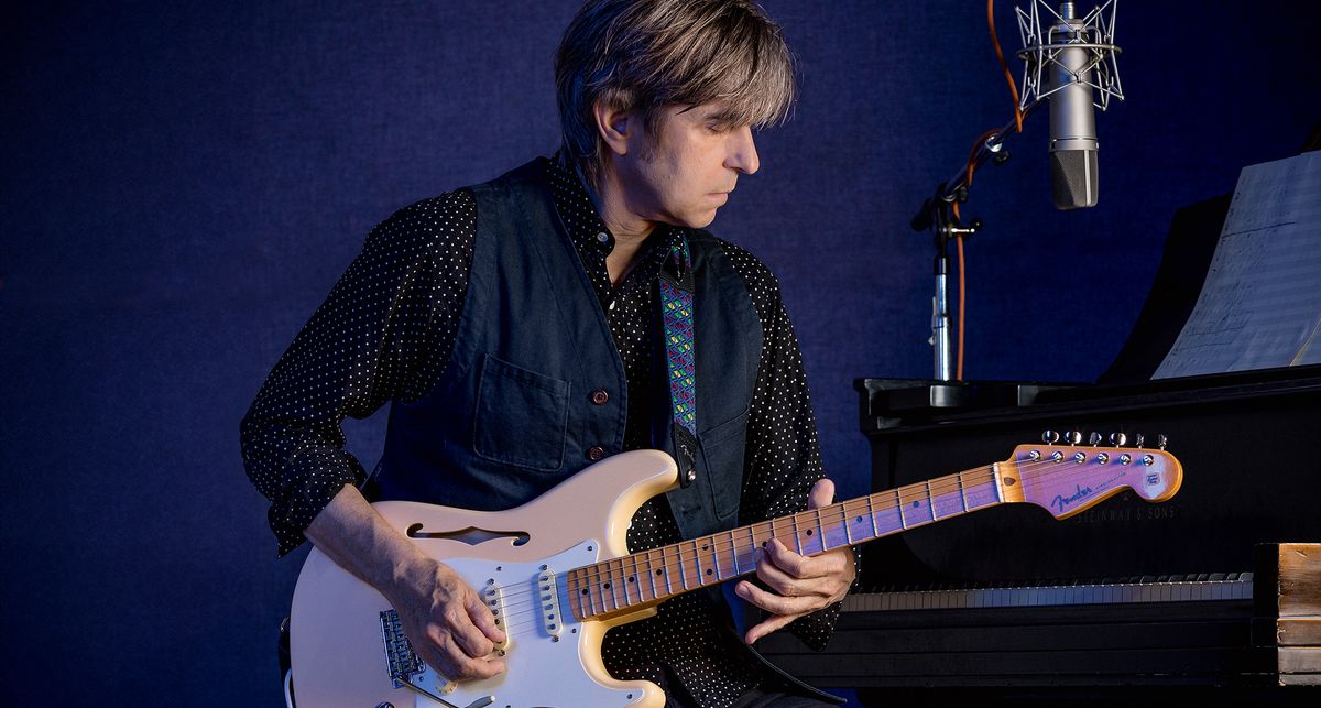 Eric Johnson sits in front of a piano with his semi-hollow Fender Stratocaster – he wears a dark shirt, and a studio mic sits on top of the piano