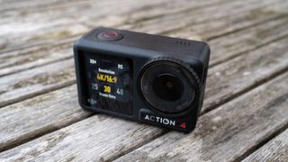 A small black camera placed upright on a faded wooden table. The left half of the camera is a simple digital screen. On the right side of the camera is the round lens and beneath that is the 'ACTION 4' branding.