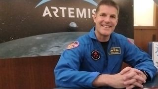 jeremy hansen smiling in his flight suit in front of an artemis 2 poster illustrating the moon and earth. the words artemis are behind his head