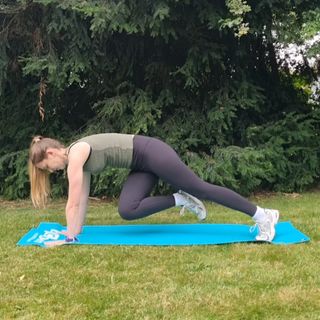 Fitness editor Ruth Gaukrodger performs a twisted mountain climber in an outdoor setting on a yoga mat. Both her arms are straight, hands touching the ground, and her left leg is extended behind her also on the ground. Her right knee is lifted and bent, so that it's close to her torso on the left side. Behind her we see a pine tree and grass.