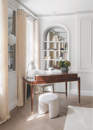 A mid-century desk in the corner of a white room with a small cream footstool.