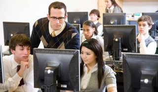 Male teacher and several teenaged students work at desktop computers.