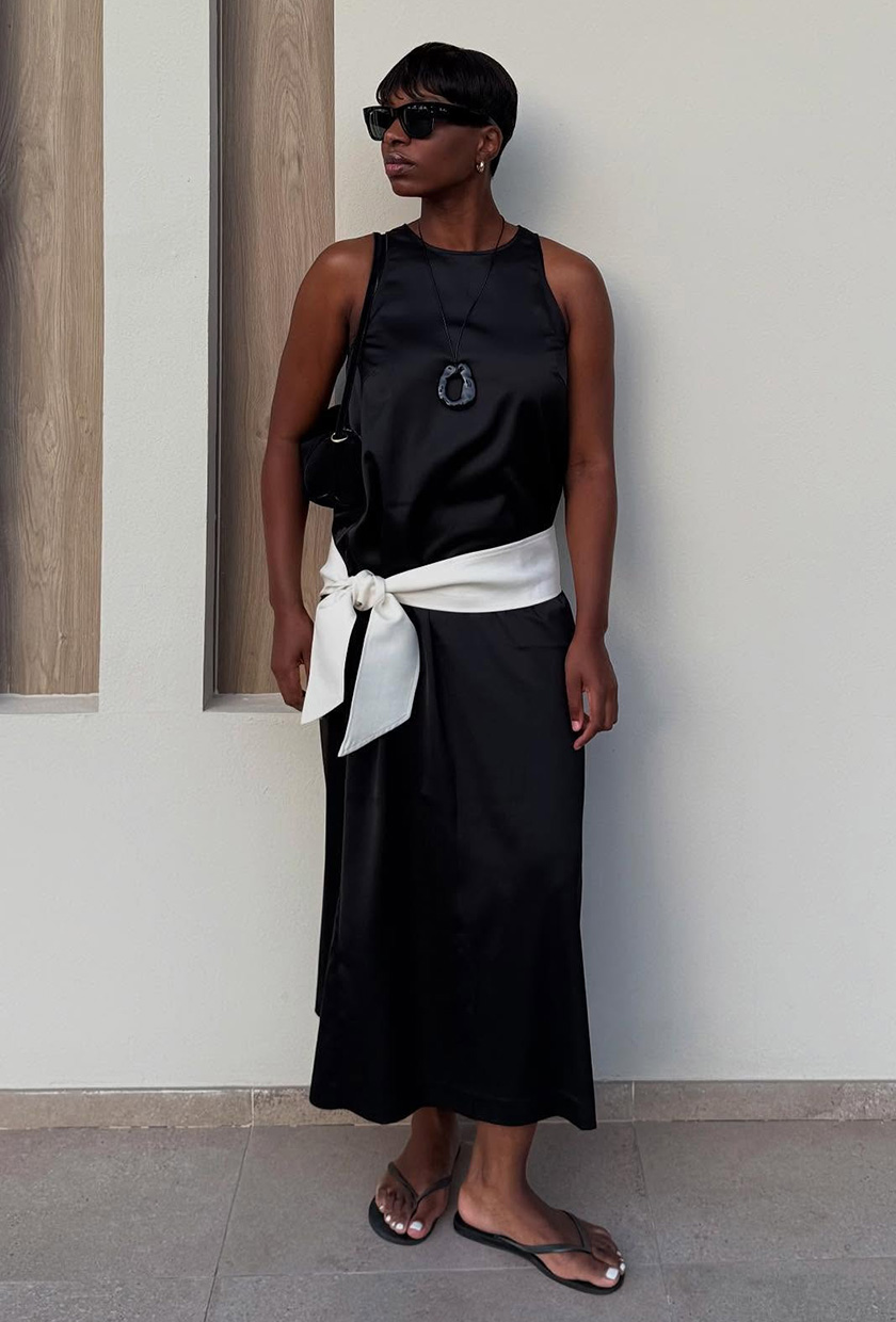 A woman in Paris wearing a black silk maxi dress styled with a long necklace, a white silk scarf tied around the waist, a shoulder bag, and flip-flops.