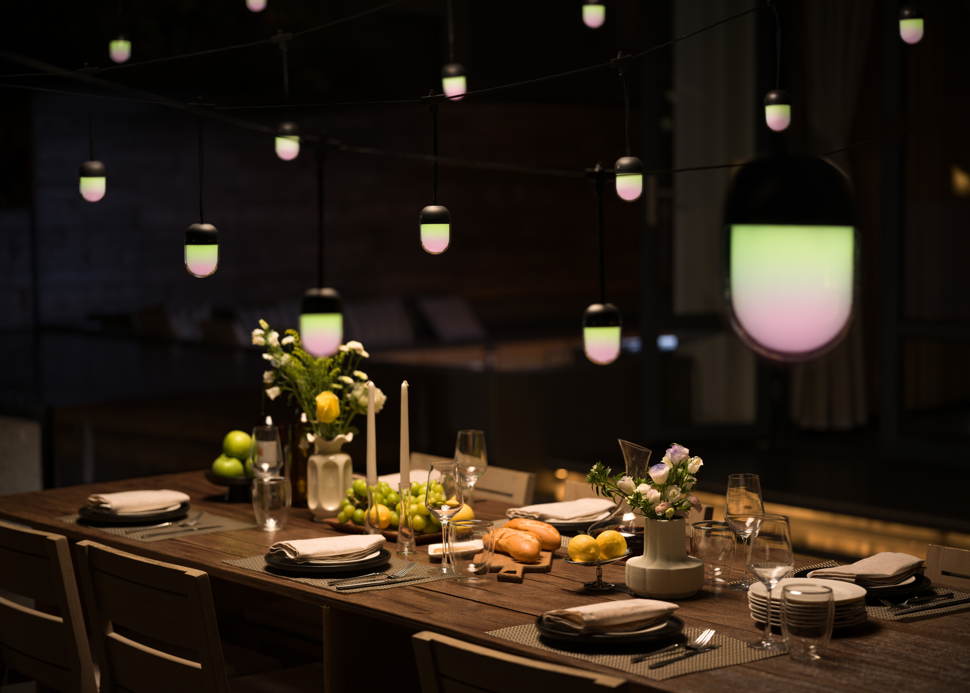 An outdoor dining area with a set dining table with flowers, candles, placemats, cutlery, crockery, and string lights with green and pink gradients