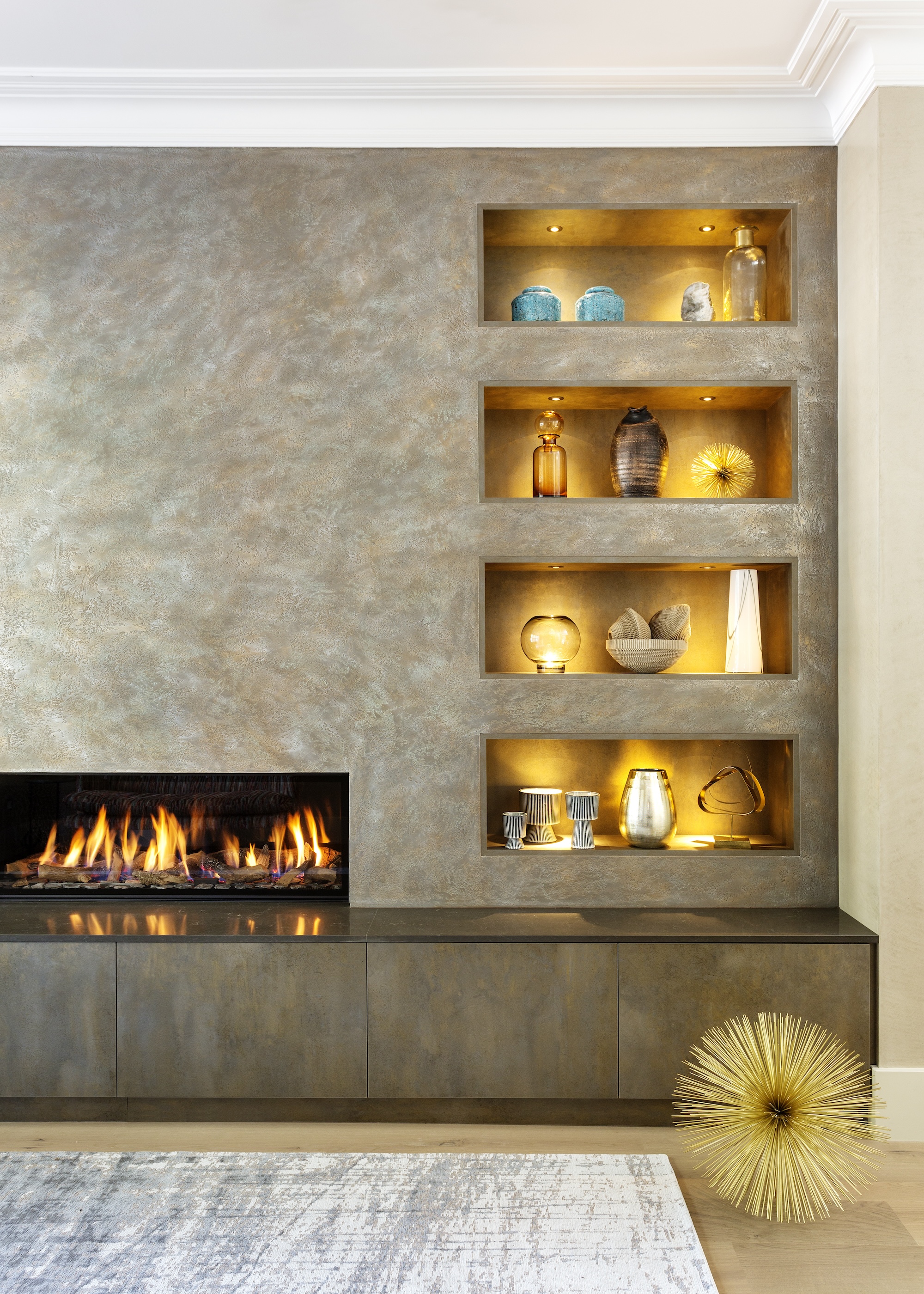 A living room with a shelf that has accent lighting around it. On the shelf are decor pieces and beside the shelf is a fireplace that has been lit up.