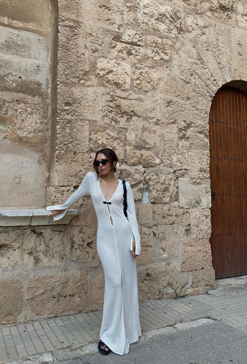 A woman's white dress outfit with a white sheer knit maxi with black sandals and a black shoulder bag.