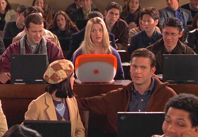 A blonde student in the middle of a crowded classroom sitting behind a distinctive orange Apple iBook