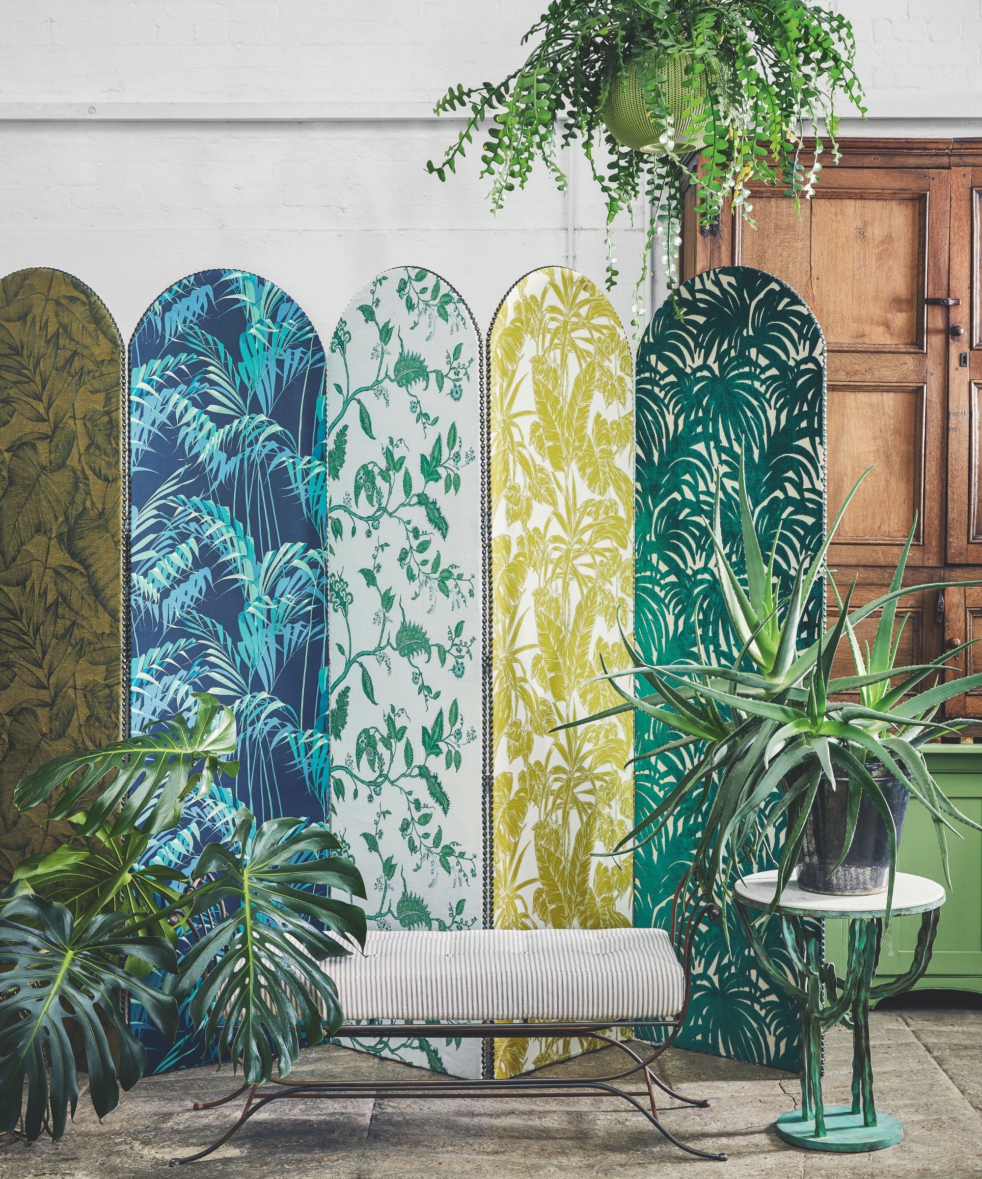 Patterned colorful room divider with houseplants against a white wall, with tiled floor, striped seating, and small circular table.